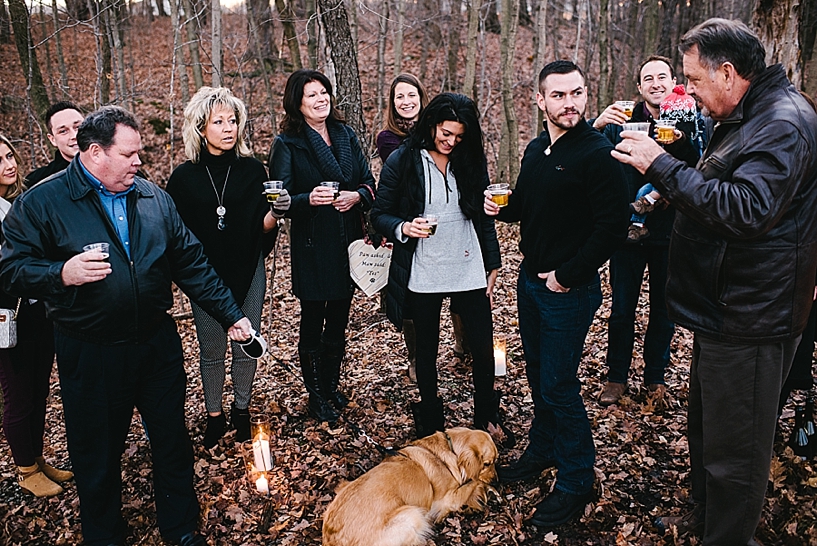 father gives champagne toast outside in woods to newly engaged couple with family gathered around
