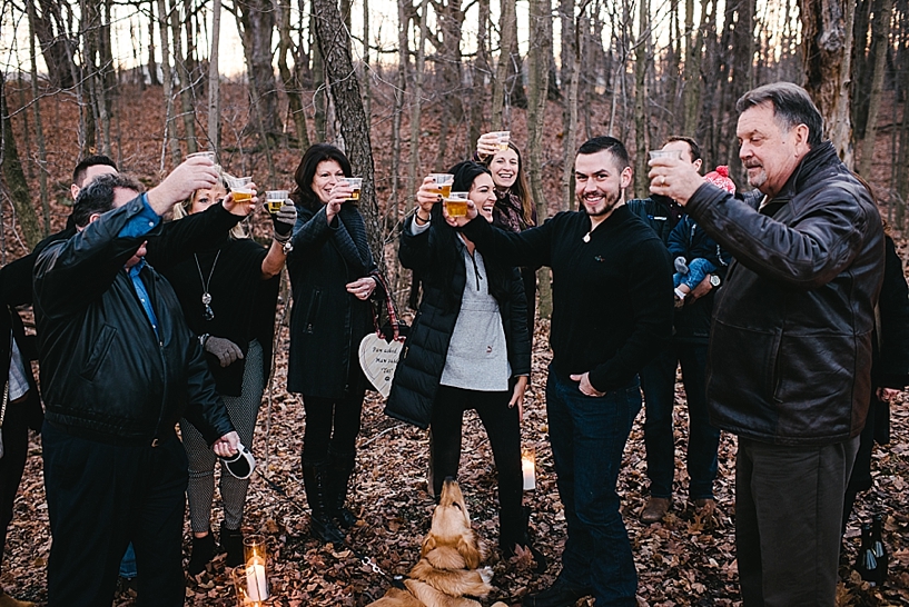 father gives champagne toast outside in woods to newly engaged couple with family gathered around