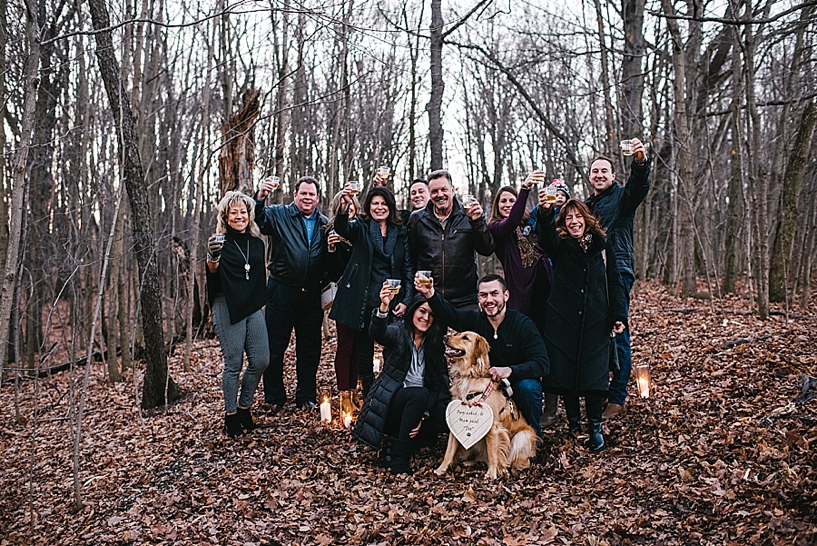 group of family members gathered around newly engaged couple toasting champagne in woods