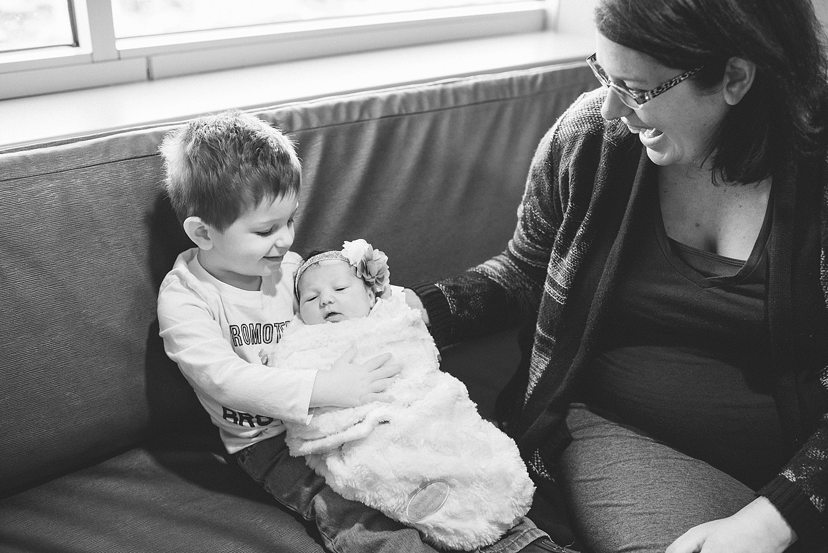 toddler boy holding newborn baby sister on his lap with mom next to him laughing