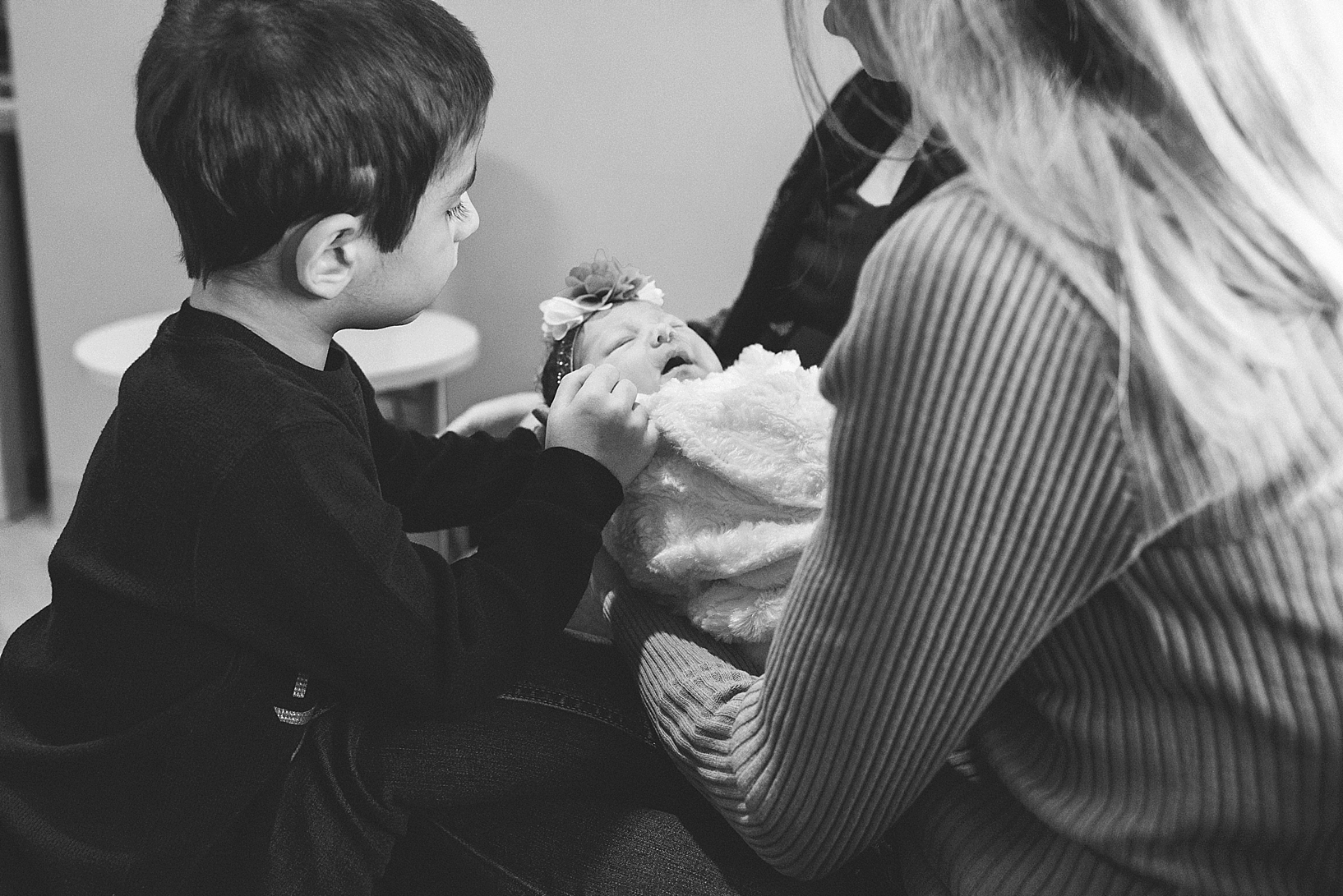 toddler boy looking at newborn baby sister wrapped in blanket in grandma's arms