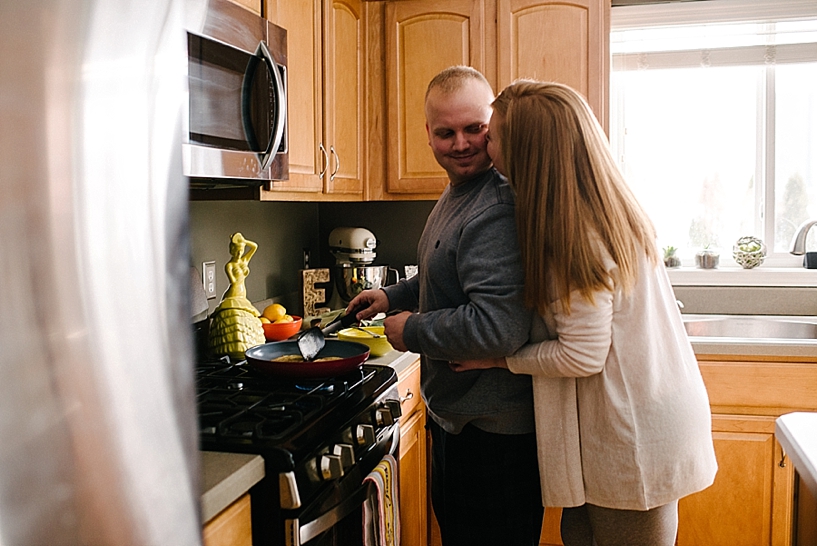 redhead woman hugs husband from behind as he makes breakfast at stove