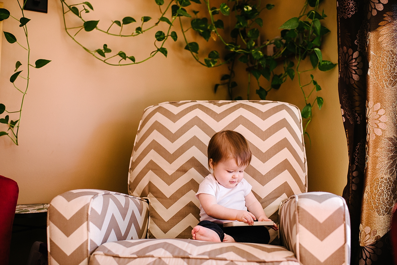 toddler in white onesie sitting on chevron chair reading a book with plant hanging on wall behind chair