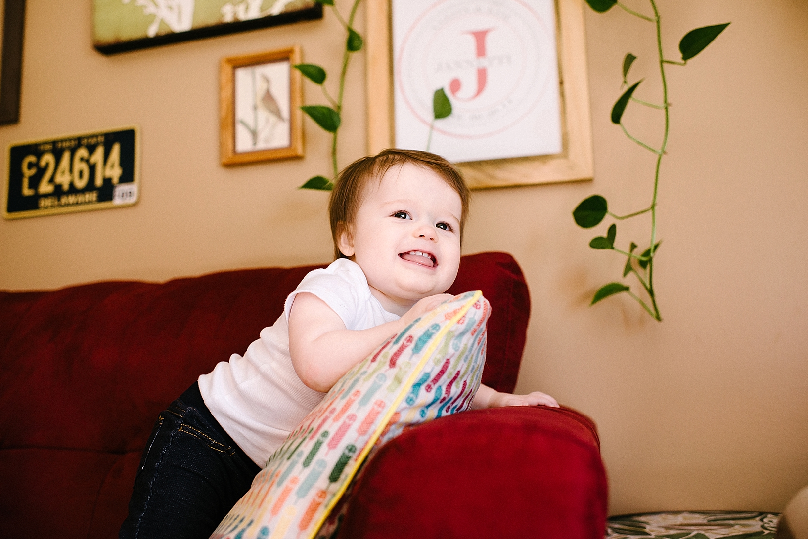 toddler boy in white onesie and denim standing on red couch smiling