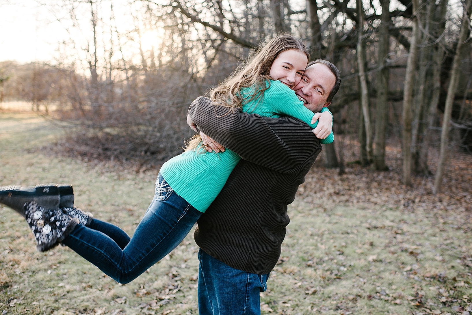 dad in brown shirt and jeans picking up teen daughter in teal shirt and hugging