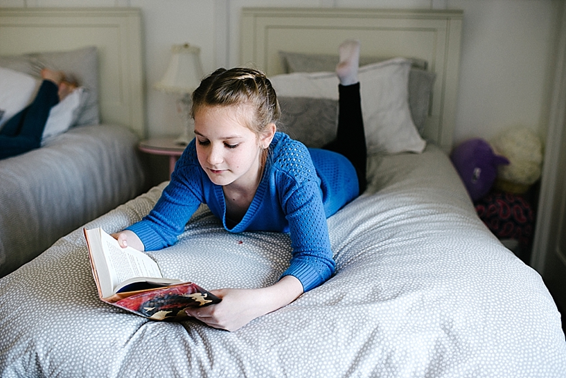 young girl in blue shirt with braided hair laying on twin bed reading book