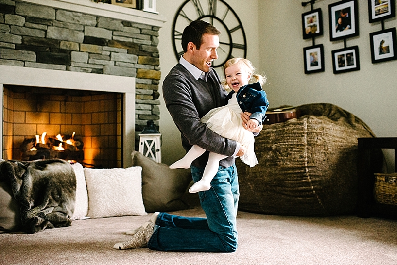 dad sitting on living room floor in front of fireplace holding laughing toddler daughter with blonde pigtails