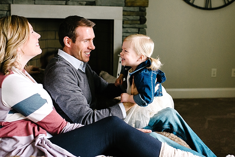 parents sitting in front of fireplace holding toddler daughter with blonde pigtails in their lap
