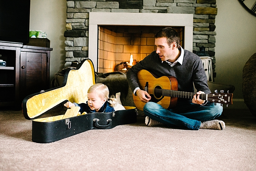 toddler girl laying in guitar case next while dad plays acoustic guitar next to her in front of fireplace