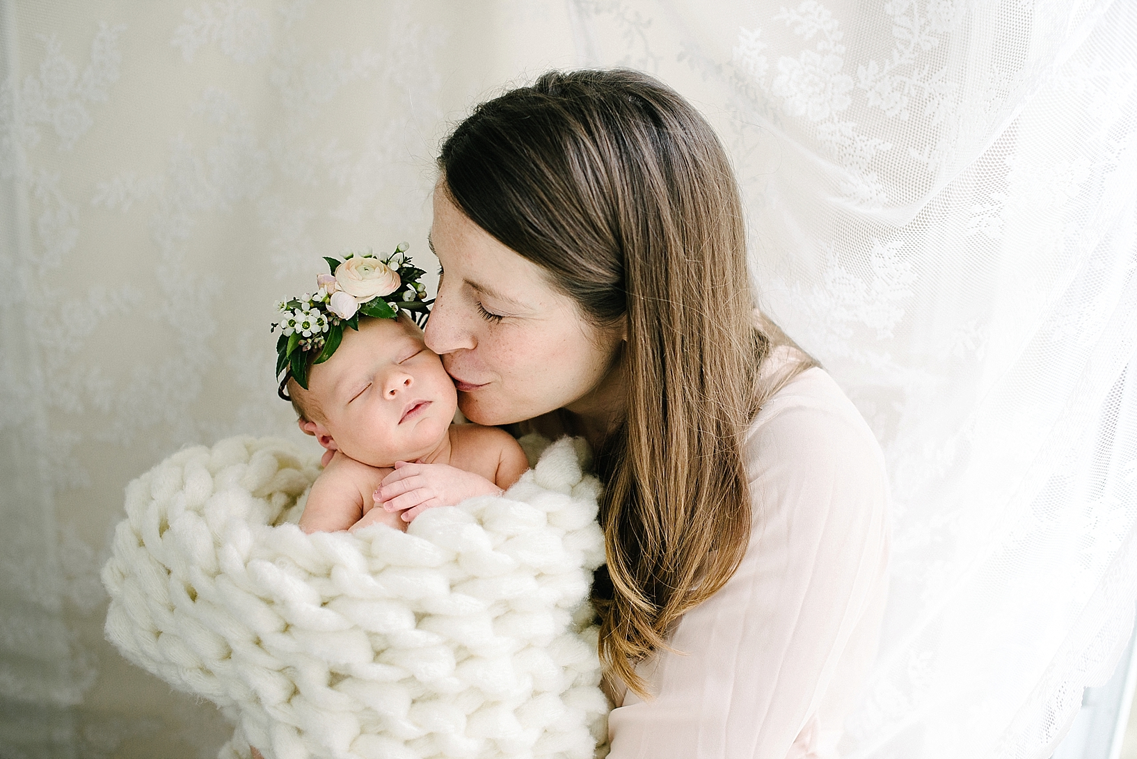 woman kissing newborn baby girl wearing floral crown wrapped in cream knit blanket