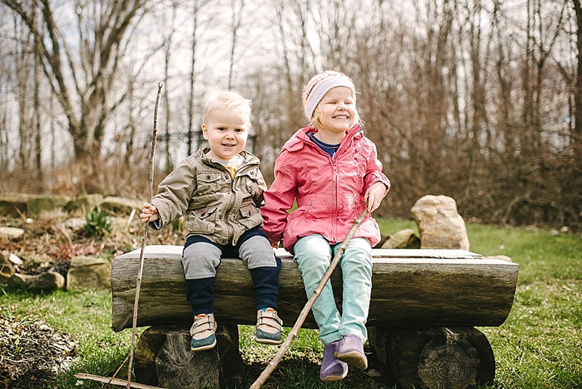 young brother and sister sitting on log bench holding sticks