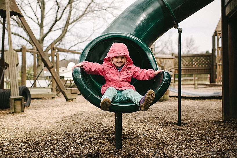 girl in pink coat and teal pants coming down green slide on playground