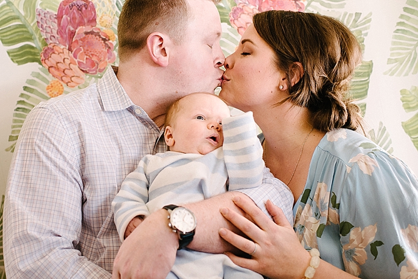 couple holding newborn baby kissing in front of tropical wallpaper