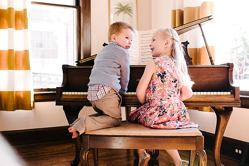 young boy in blue sweater and little girl in floral print dress sitting on bench playing piano