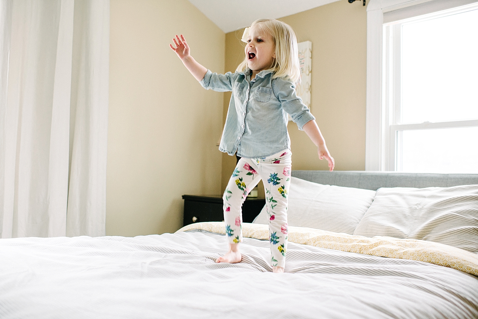 blonde toddler girl wearing denim shirt and floral leggings jumping on bed