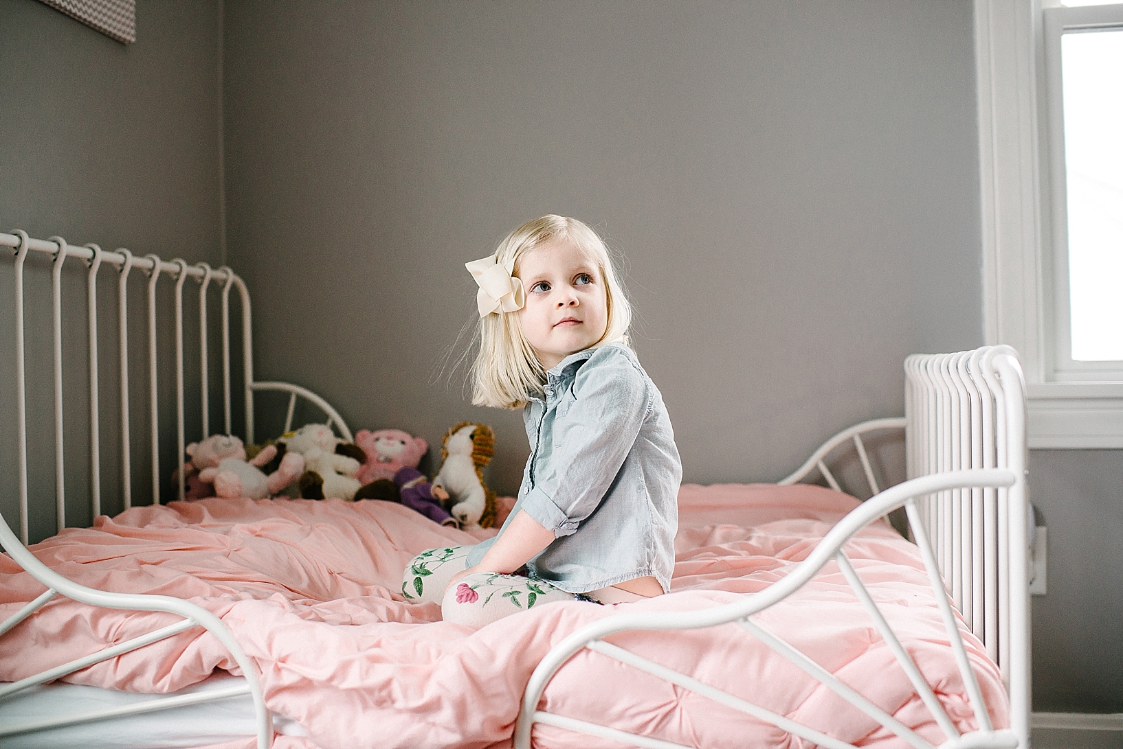 blonde toddler girl sitting on white wrought iron bed with pink comforter
