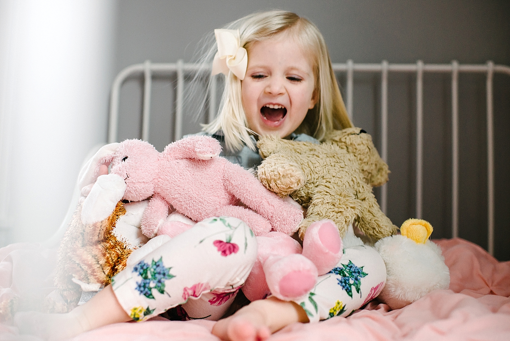 blonde toddler girl wearing floral leggings sitting on white wrought iron bed with stuffed animals in her lap