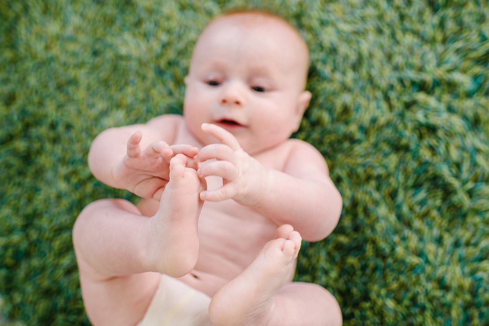 infant laying on green carpet playing with his feet
