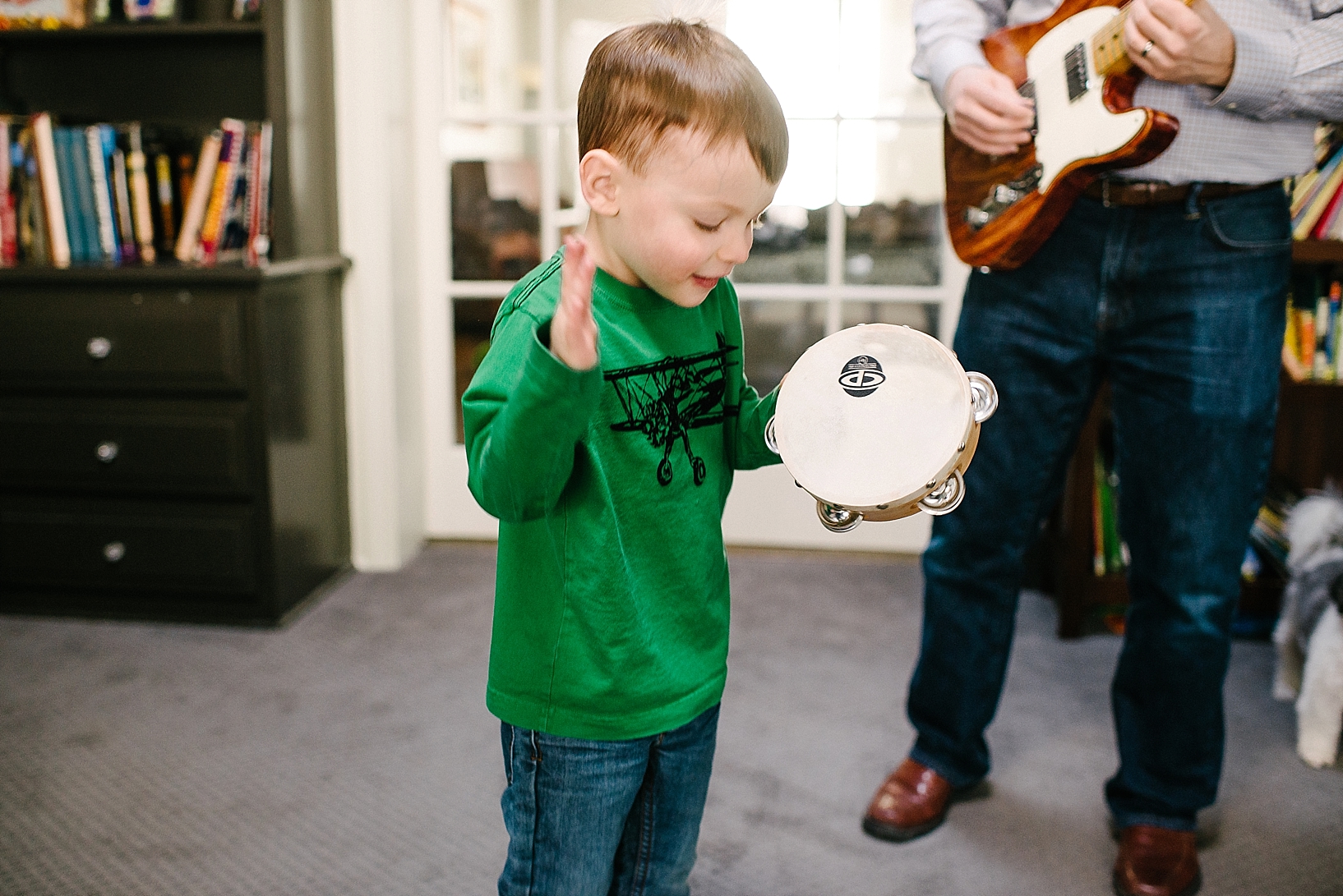 little boy playing tambourine with his dad playing electric guitar in living room