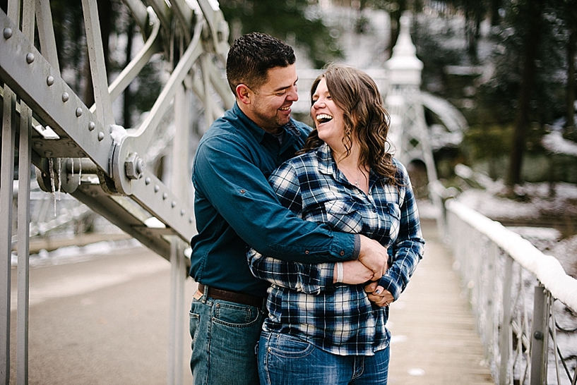 man hugging woman in blue plaid shirt from behind and laughing on silver bridge