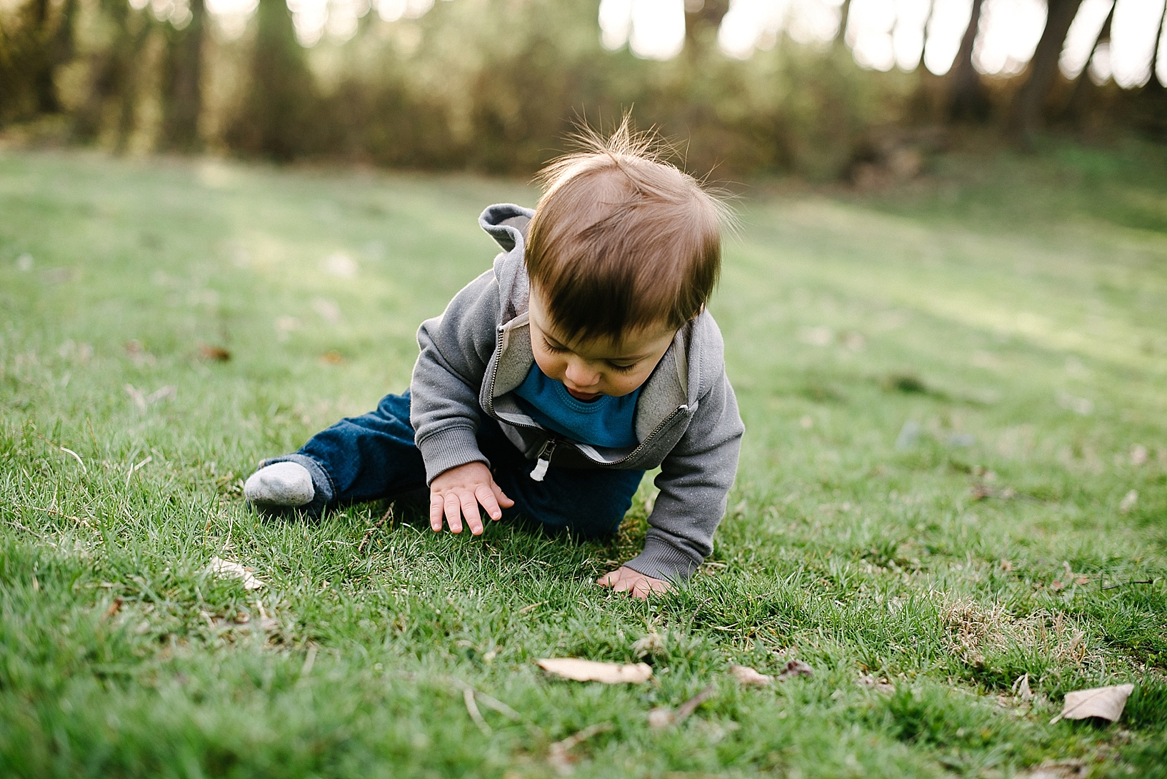 one year old boy wearing grey hoodie and jeans sitting on grassy hill