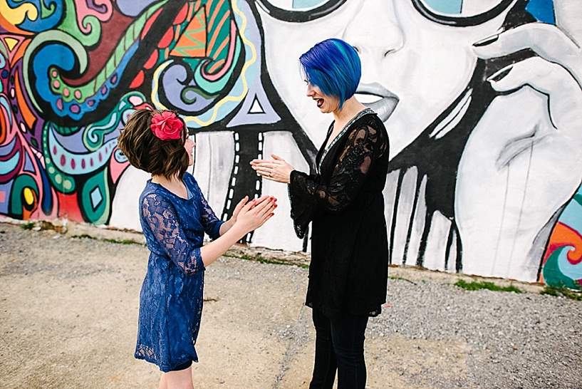 woman with blue hair playing clapping game with young daughter wearing blue lace dress in front of colorful wall mural