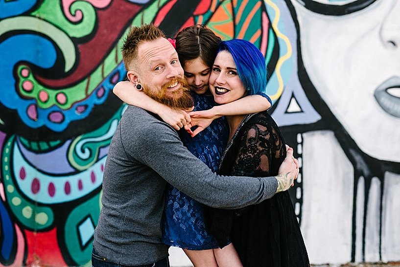 man with beard and piercings hugging wife with blue hair and young daughter standing in front of graffiti mural