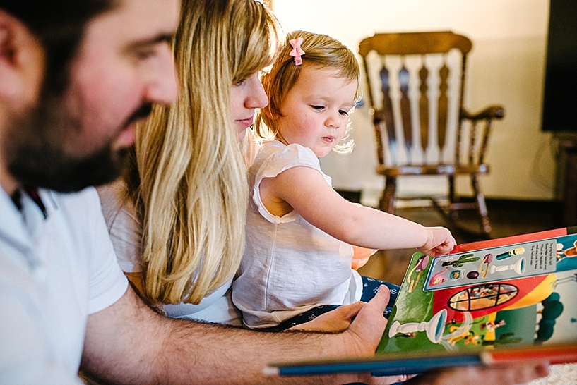 toddler girl reading book with her parents in living room