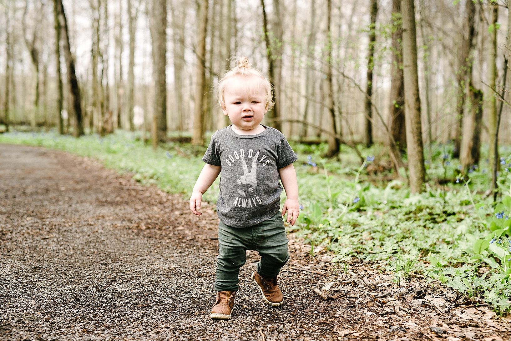 hipster toddler boy wearing man bun and Good Vibes tshirt walking through woods in spring