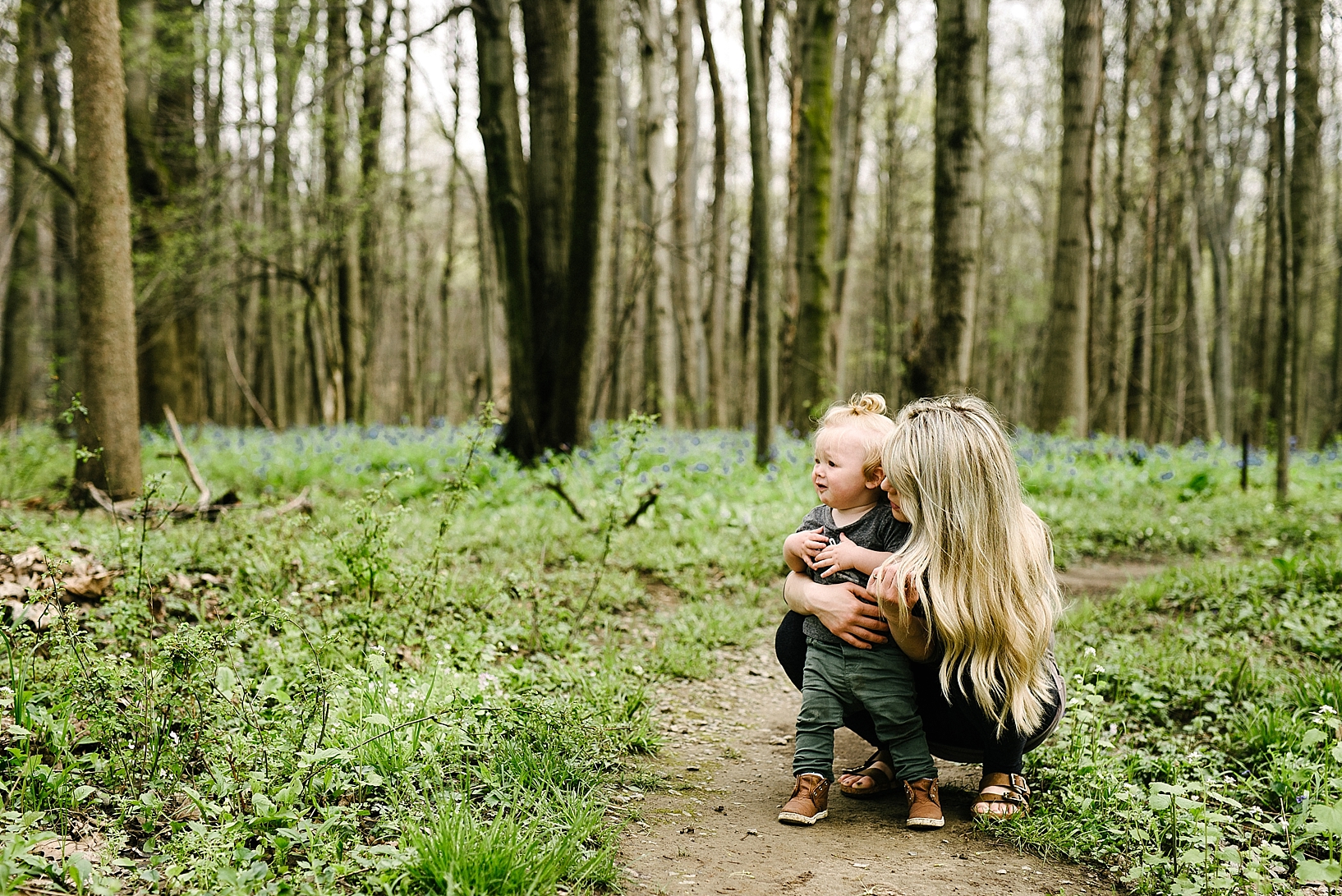 woman with long wavy blonde hair kneeling by son in woods along trail of bluebells