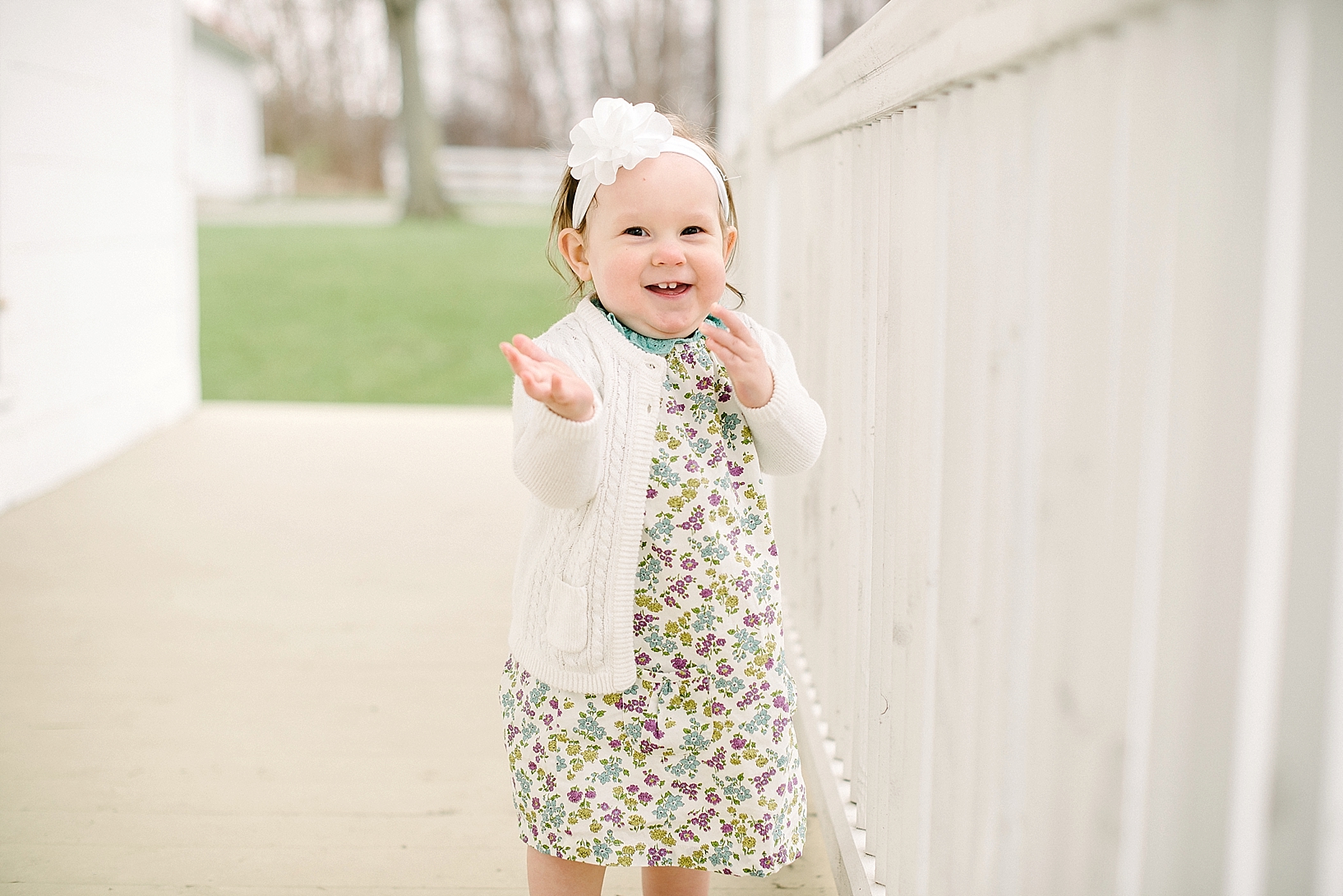 one year old girl wearing floral print dress and cream cardigan standing on front porch
