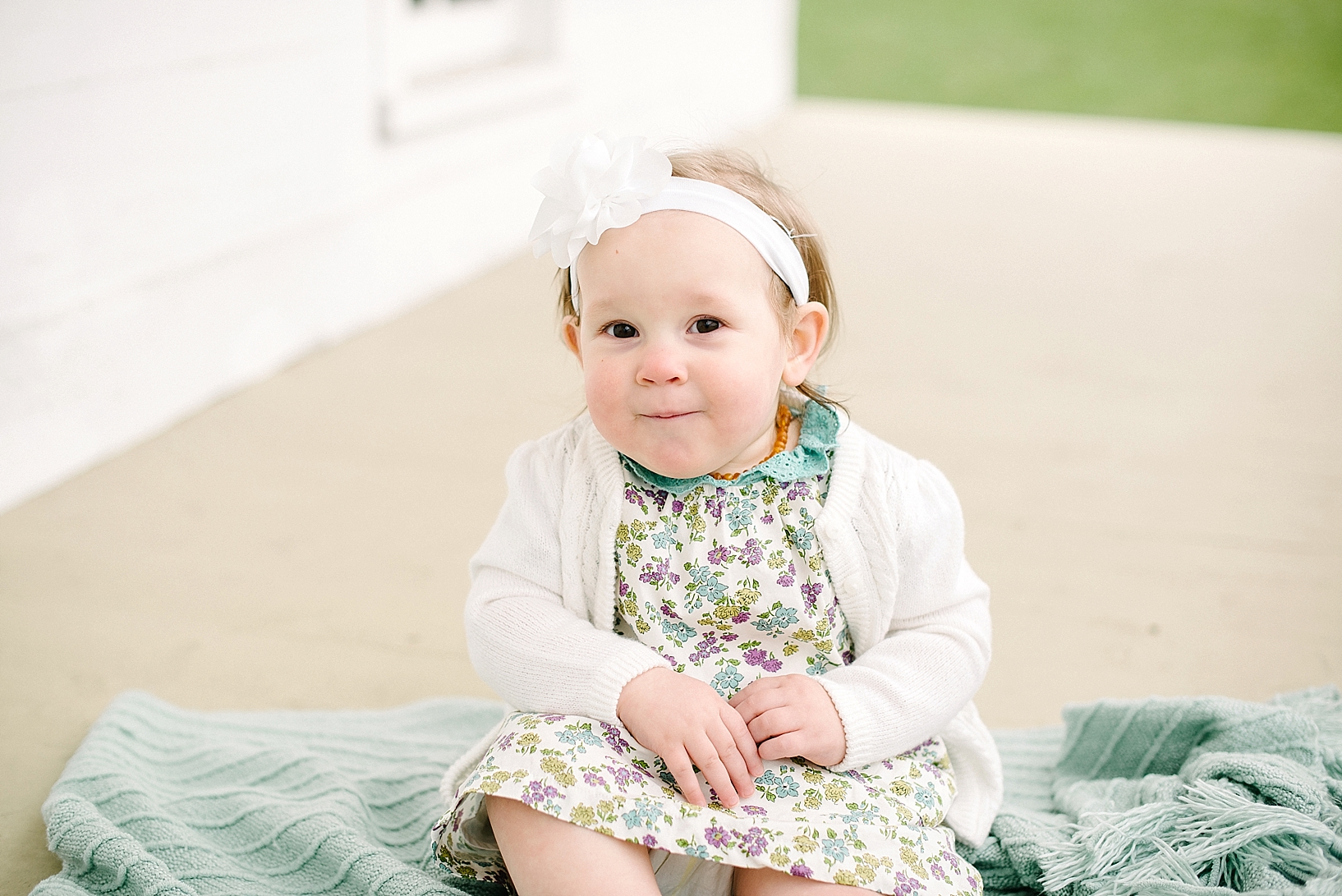 one year old girl wearing floral print dress and cream cardigan sitting on teal blanket on porch