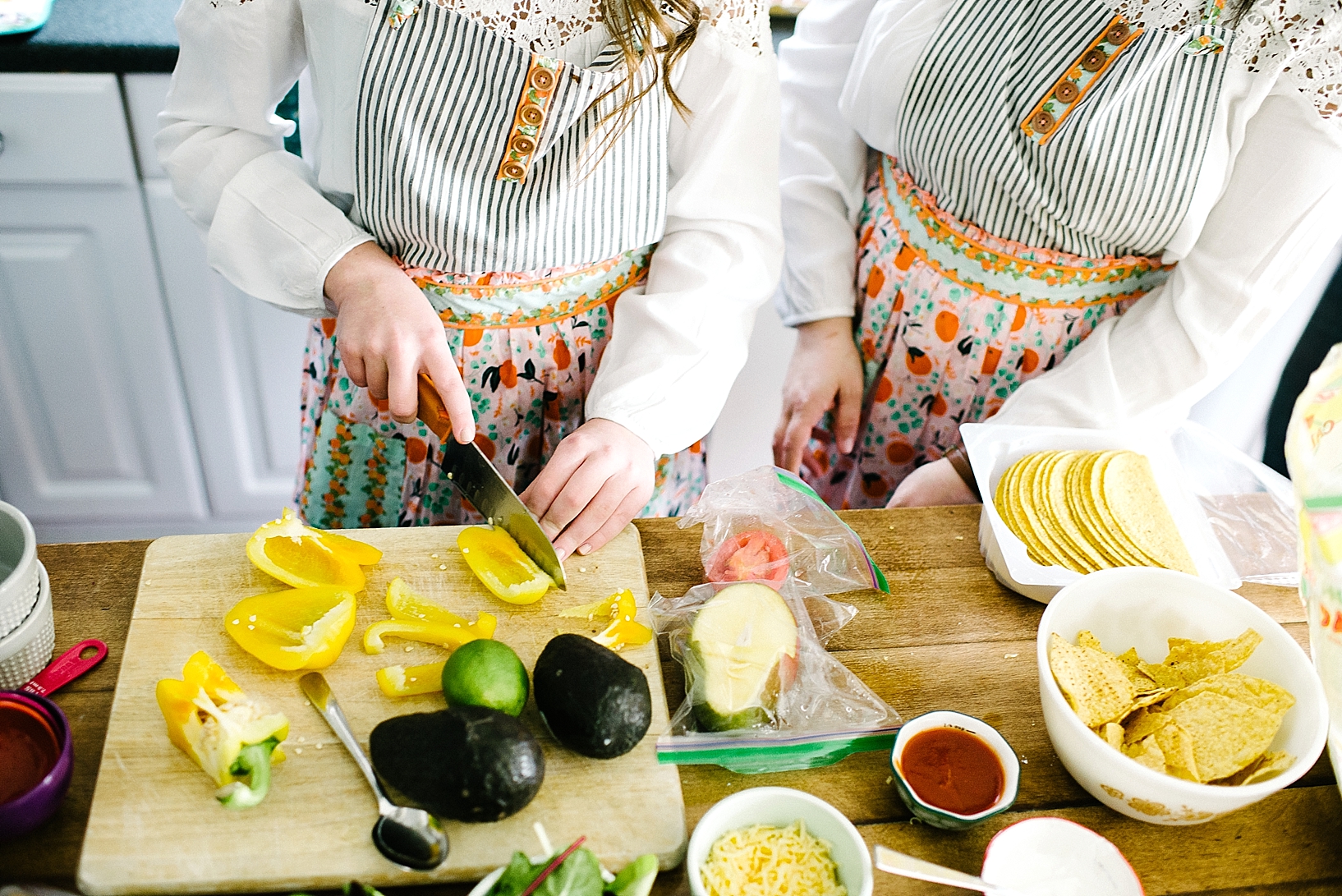 mother and teen daughter wearing patterned aprons standing at kitchen island chopping yellow peppers