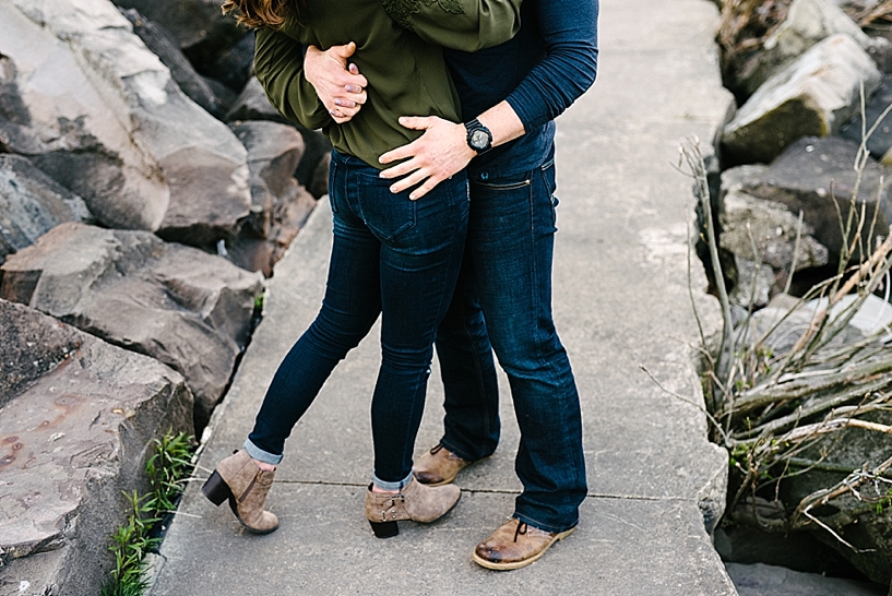 man wearing navy shirt and jeans hugging woman wearing olive green shirt and jeans on walking trail lined with rocks