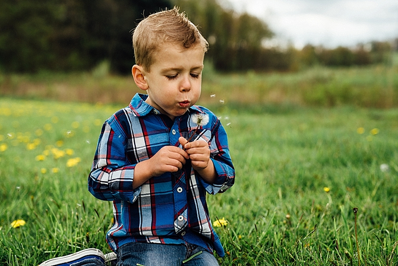 young boy wearing blue converse and blue and red plaid shirt sitting in grass blowing dandelion