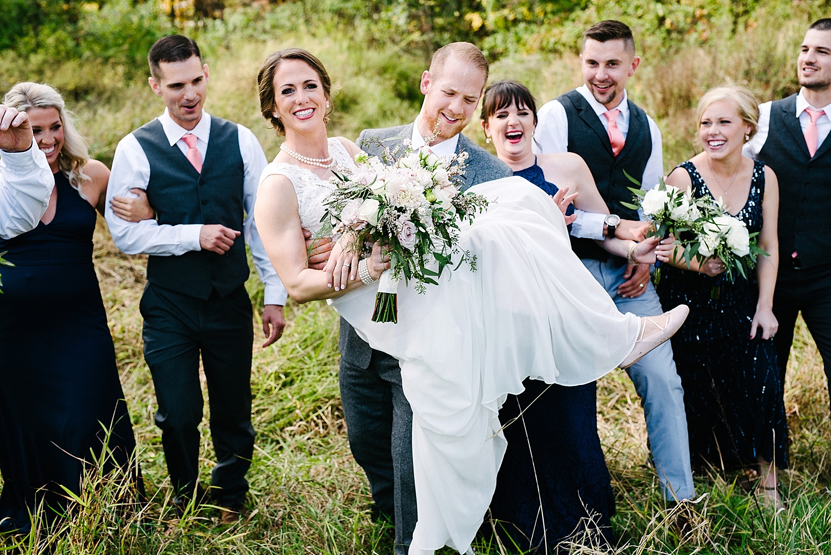 groom carrying bride out of field of grass with bridal party behind him