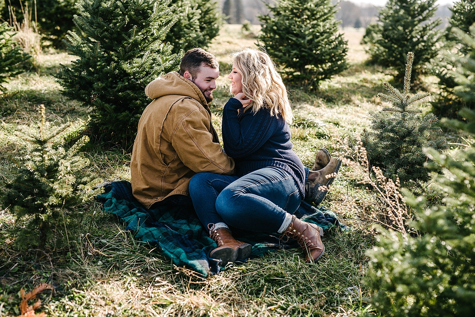 couple sitting on plaid blanket among pine trees at tree farm