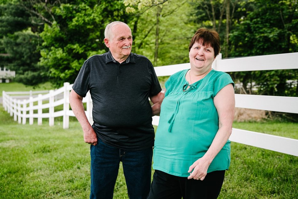 Grandma and grandpa smile and share a look by white picket fence