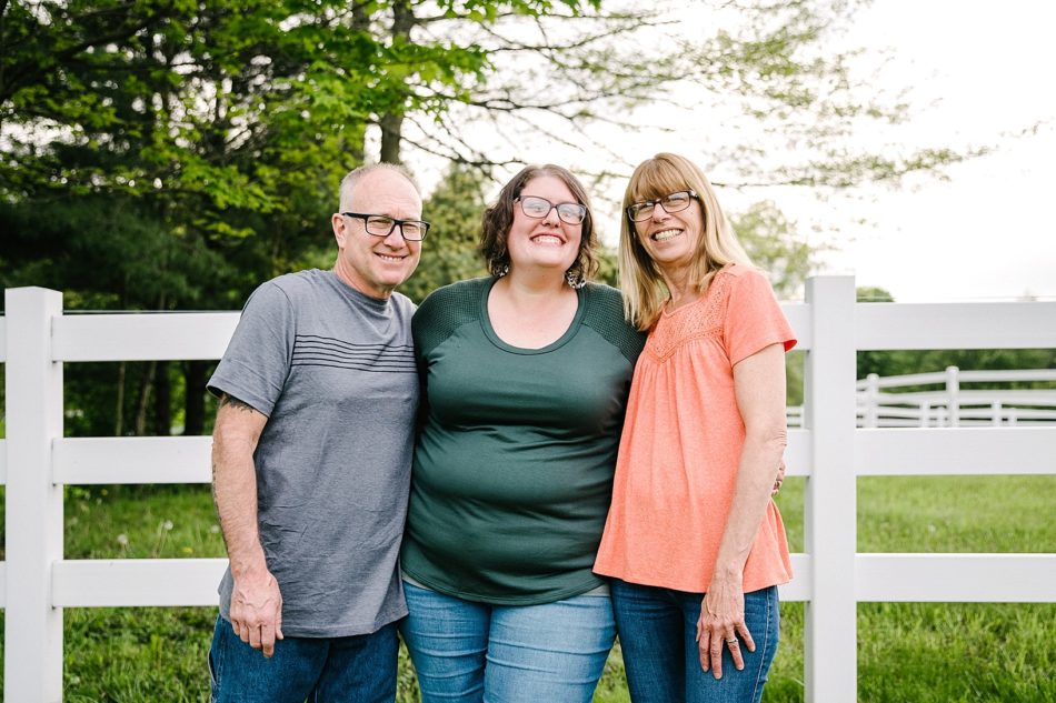 Three family members smile and hug by white picket fence in generational family session