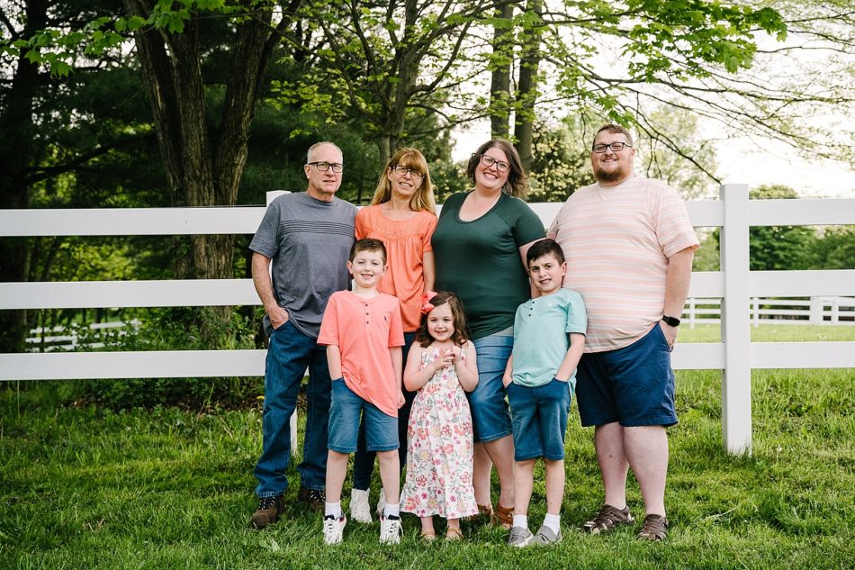 A whole family of 7 smiles by a white picket fence in generational family session