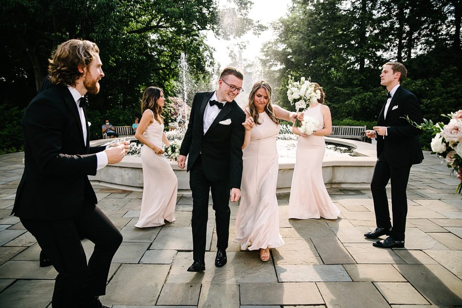 Groomsman and bridesmaid walk down aisle of wedding party in front of fountain during Fellows Riverside Gardens wedding