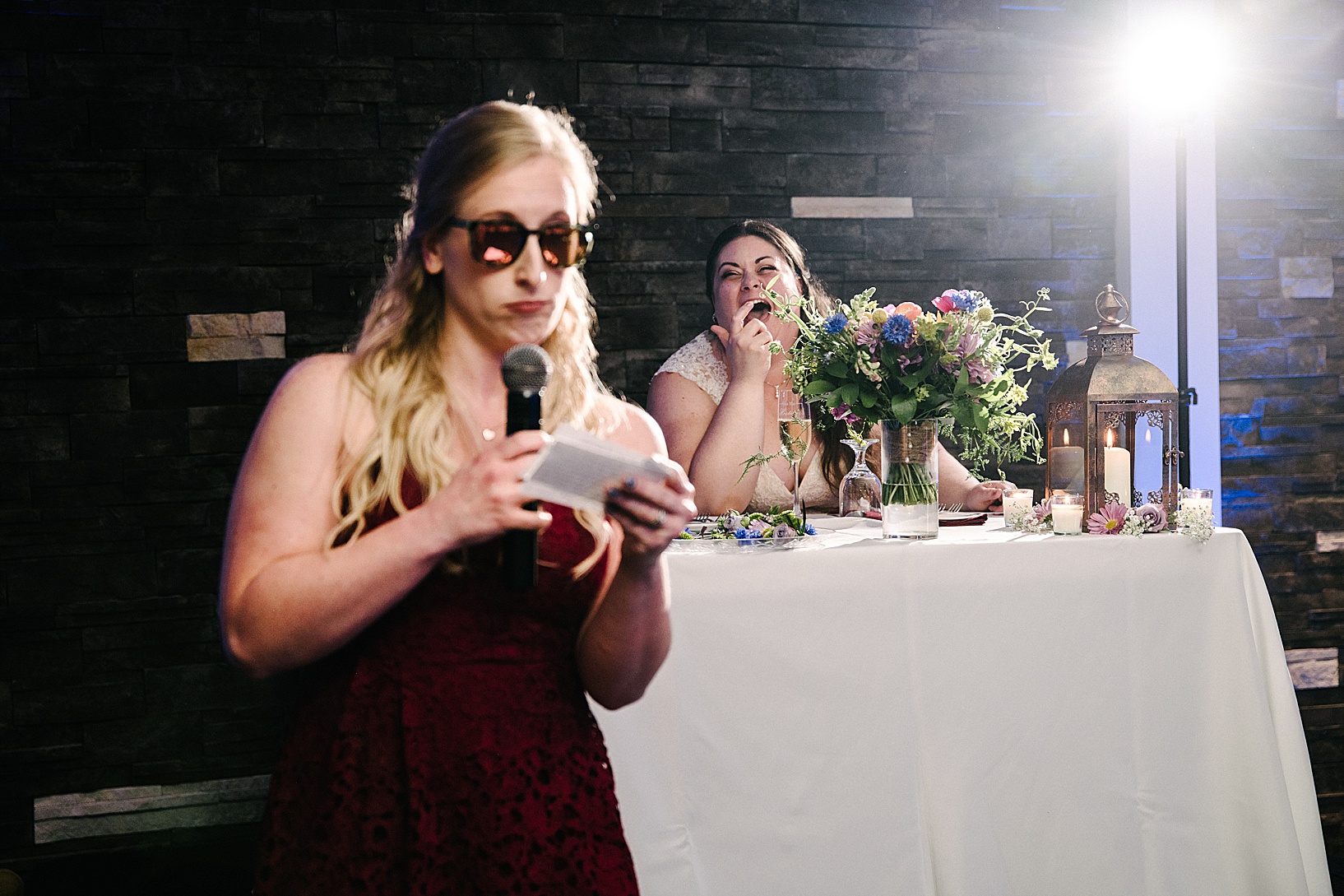 Bride laughing in background as bridesmaid gives her speech wearing sunglasses