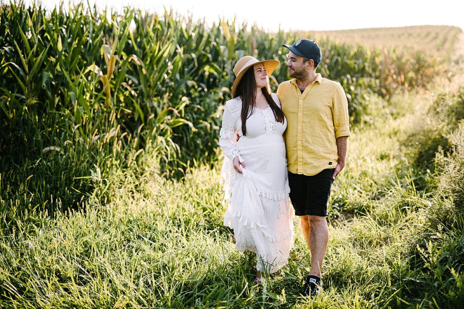 Man in yellow shirt and his pregnant wife in a white gown and sun hat walk through a field with arms around each other.