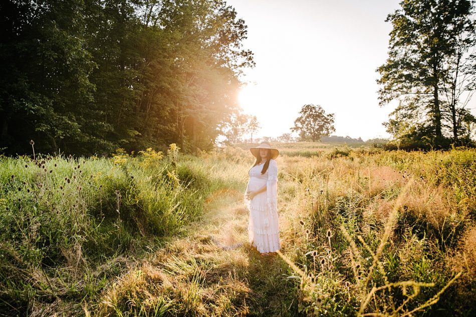 Pregnant woman stands in field holding her belly, with the sun glowing through trees behind her.