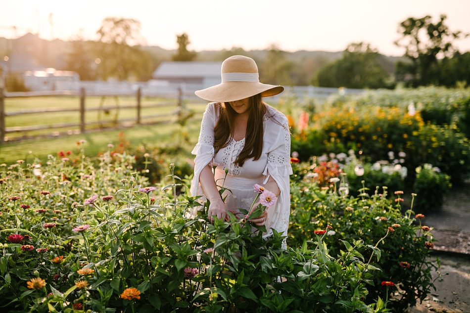 Pregnant woman picks pink wildflowers in white gown and floppy sun hat