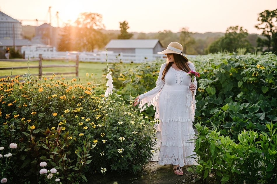 Pregnant woman in white gown and sun hat walks through rows of wildflowers