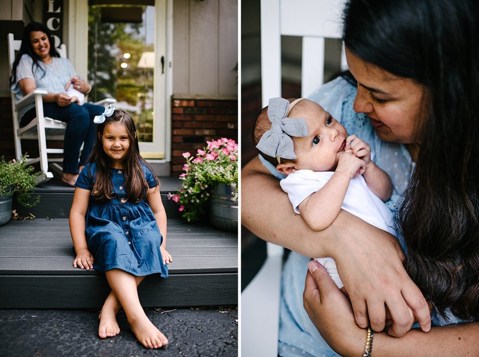 Young girl in denim dress sits on edge of family front porch with mom in the background holding a baby girl in white rocking chair.