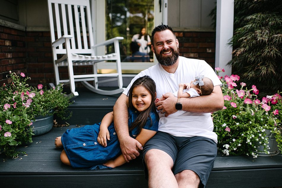 Dad holds baby daughter and hugs older daughter while sitting on family front porch