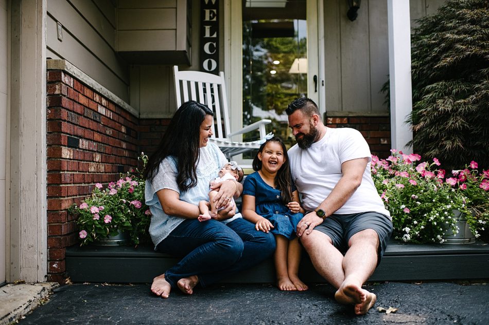Mom holding baby, father, and older daughter smile while sitting on family's front porch.