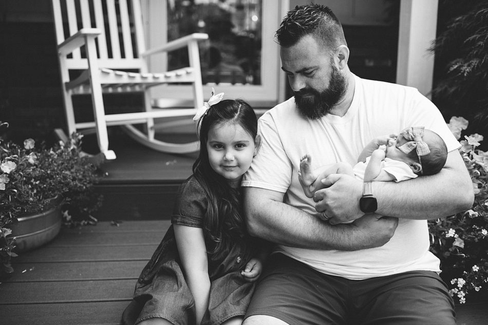 Father holds baby daughter and smiles at older daughter sitting next to him on their front porch